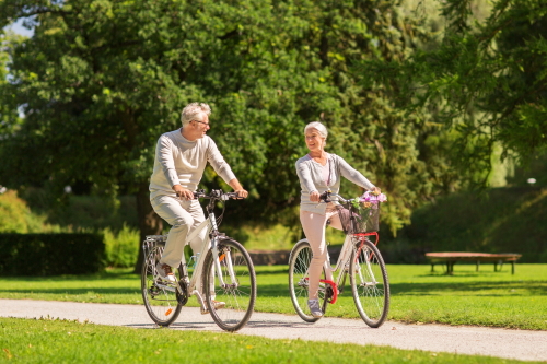 This image: an elderly couple ride bikes through a green park, whilst looking and smiling at each other. The map: the map zooms into Littlebourne, showing key bus routes and stops in blue, proposed and existing footpaths in orange, pink and blue, and local facilities indicated by gold map markers. The user can click on the gold markers to find out approximate walking times from the proposed site entrance.